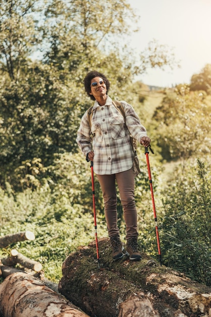 Mujer negra madura con mochila y bastones de trekking disfrutando de la vista del bosque mientras camina por la naturaleza.