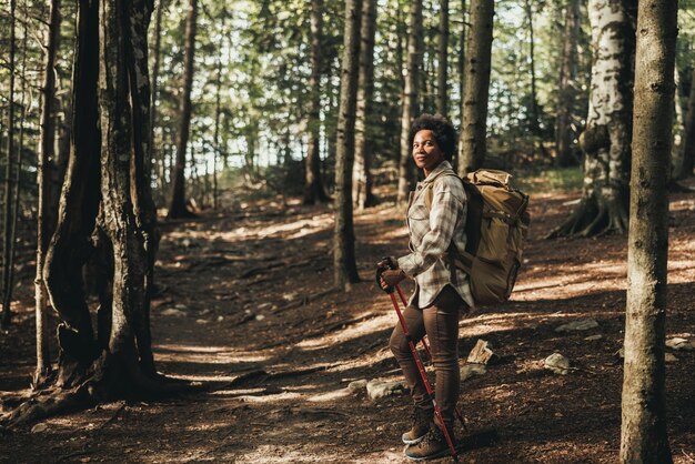 Mujer negra madura parada sola afuera y cargando una mochila mientras camina con bastones de trekking en la montaña.