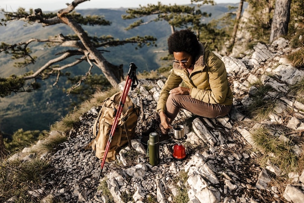 Mujer negra madura sentada en la cima de una colina y tomando un descanso para el té durante su caminata en las montañas.