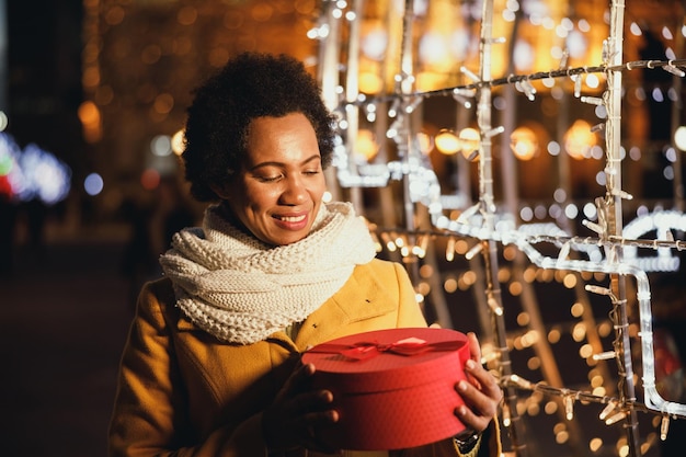 Mujer negra de mediana edad sosteniendo una caja de regalo roja mientras caminaba en el mercado navideño de invierno en la ciudad.