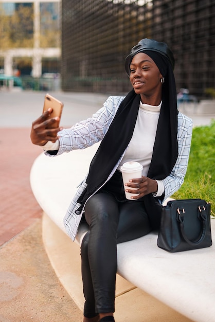 Mujer negra musulmana con estilo positivo tomando selfie en el teléfono inteligente