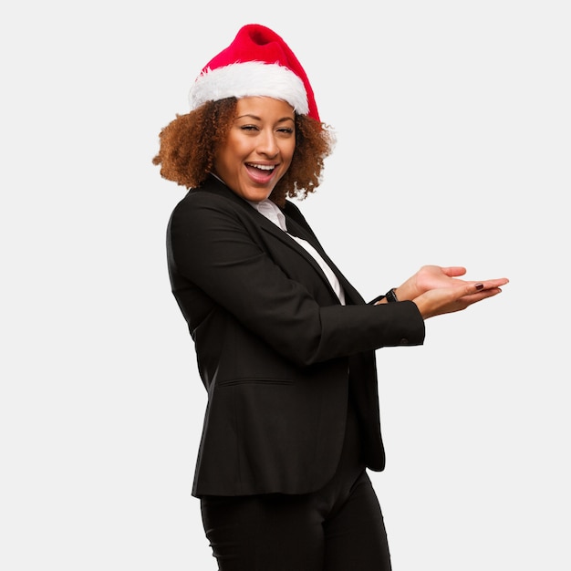 Mujer negra del negocio joven que lleva un sombrero de santa de los chirstmas que lleva a cabo algo con las manos