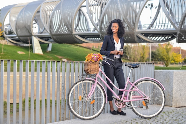 Mujer negra de negocios de pie junto a su bicicleta vintage hablando por teléfono móvil
