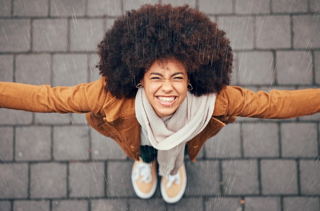 Mujer negra de pie para sentir la lluvia y feliz en la ciudad en invierno con una sonrisa afro o moda de moda Mujer al aire libre arriba o felicidad en salpicaduras de llovizna y cara en metro urbano o ciudad con cabello natural