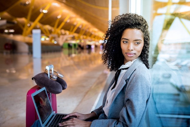 Mujer negra que trabaja con la computadora portátil en el aeropuerto esperando en la ventana