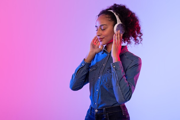 Una mujer negra relajada disfrutando de la música con auriculares, vestido de denim y telón de fondo colorido.