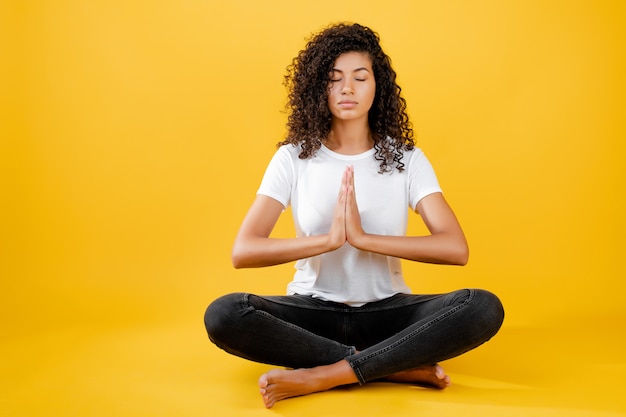 Mujer negra relajada meditando en pose de yoga aislado sobre amarillo