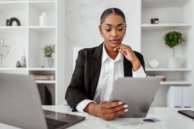 Mujer negra segura con ropa formal usando una tableta digital charlando en un gadget para negocios en la oficina