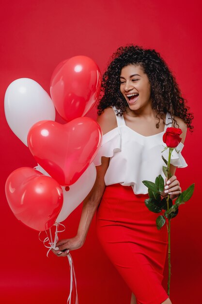 Mujer negra sonriendo con globos en forma de corazón y rosa aislado