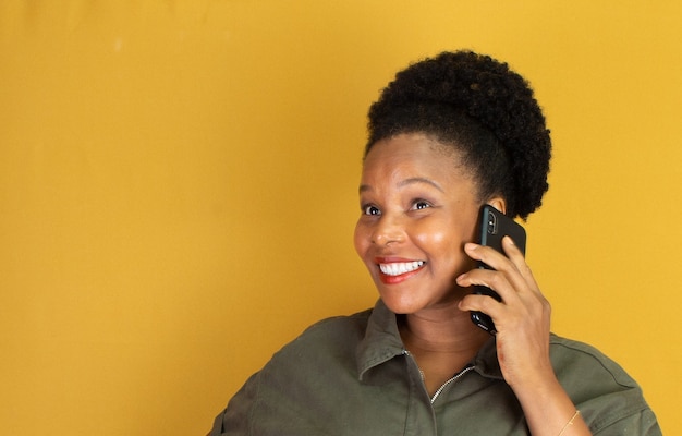 Mujer negra sonriendo con ropa verde sosteniendo un teléfono celular sobre una superficie amarilla