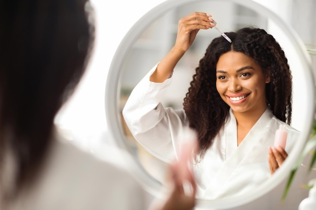 Mujer negra sonriente aplicando suero para reparar el cabello mientras está de pie cerca del espejo