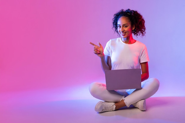 Una mujer negra sonriente con una computadora portátil sentada con las piernas cruzadas apuntando hacia un lado en el espacio libre