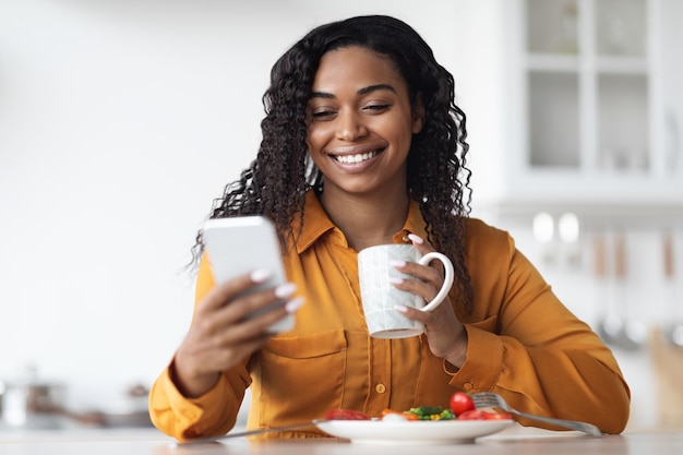 Mujer negra sonriente revisando su teléfono inteligente mientras desayuna