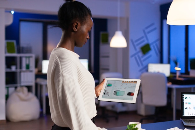 Mujer negra trabajando en la fecha límite analizando gráficos en tablet pc trabajando hasta altas horas de la noche