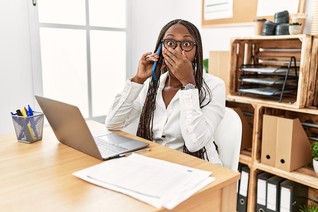 Mujer negra con trenzas trabajando en la oficina hablando por teléfono sorprendida cubriendo la boca con las manos por error. concepto secreto.