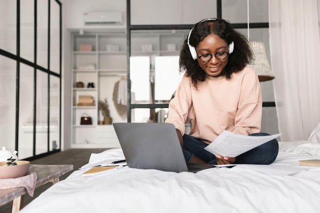 Mujer negra usando laptop y leyendo papel sentada en el dormitorio