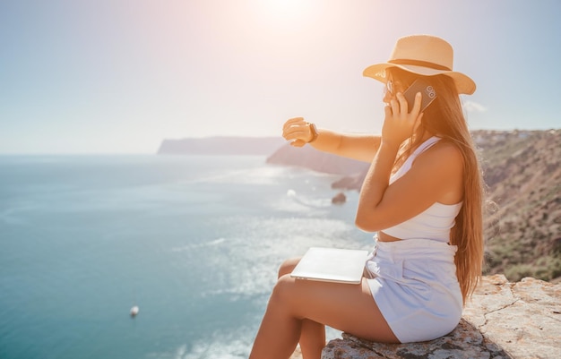 Mujer nómada digital en el sombrero una mujer de negocios con una computadora portátil se sienta en las rocas junto al mar durante