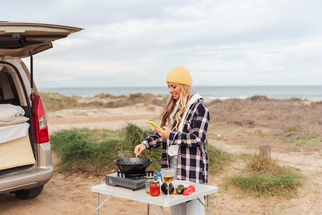 Mujer nómada que usa teléfono móvil mientras cocina fuera de su autocaravana cerca de la playa Concepto de viaje y estilo de vida nómada
