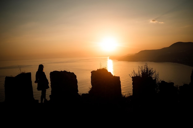 Una mujer o un hombre de pie sobre una roca y mirando directamente la puesta de sol junto al mar El concepto de naturaleza y belleza Puesta de sol naranja Silueta al atardecer