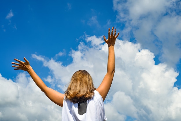 Foto una mujer o una niña se para y extiende sus manos hacia el cielo. la niña levantó las manos contra el cielo azul.