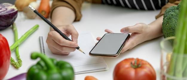 Mujer observando el ingrediente en el cuaderno mientras busca información en la pantalla en blanco del teléfono inteligente en la mesa de la cocina con verduras frescas