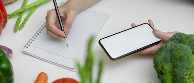 Mujer observando el plan de dieta mientras busca información en la pantalla en blanco del teléfono inteligente en la mesa de la cocina con verduras frescas