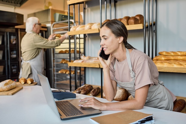 Mujer ocupada en delantal usando laptop y llamando a clientes