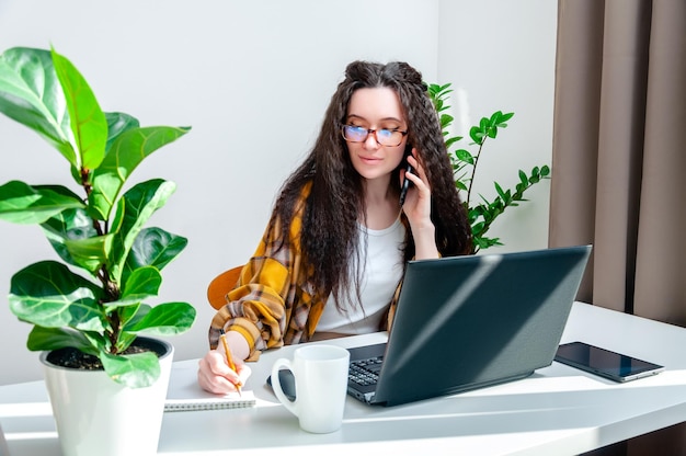 Mujer ocupada con gafas hablando por teléfono y escribiendo en un cuaderno en una mujer en el lugar de trabajo que trabaja en