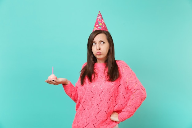Mujer ofendida en suéter rosa de punto, sombrero de cumpleaños mirando hacia arriba, sostenga la torta en la mano con vela aislada en el retrato de estudio de fondo de pared azul turquesa. Concepto de estilo de vida de personas. Simulacros de espacio de copia.