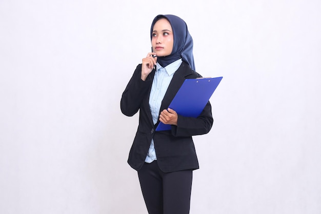 Mujer de oficina madura asiática en hijab de pie pensando a la derecha llevando un bolígrafo y abrazando un clipb