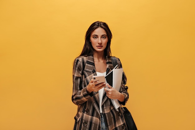 Mujer de ojos verdes con una chaqueta elegante de gran tamaño mira a la cámara Una chica morena de pelo largo sostiene cuadernos y teléfono en un fondo amarillo aislado