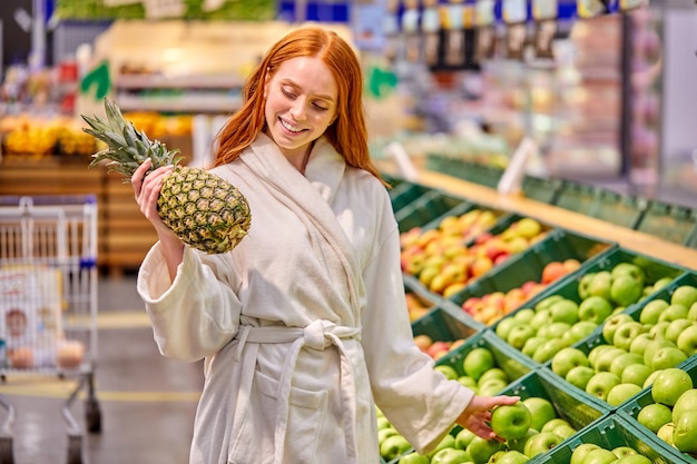 Mujer optimista comprando frutas, eligiendo piña y manzanas, vistiendo albornoz, sonriendo