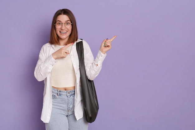 Mujer optimista positiva con una bolsa negra con camisa blanca y jeans de pie y señalando a un lado mostrando espacio de copia para publicidad posando aislado sobre fondo púrpura