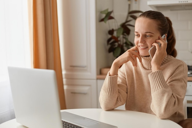 Mujer optimista positiva con cabello oscuro usando suéter beige sentada en la cocina y trabajando en una laptop mirando la pantalla y hablando a través de un teléfono inteligente con una sonrisa