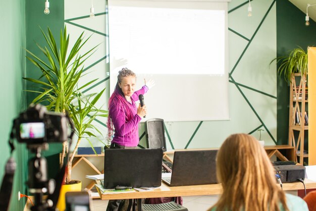 Mujer oradora de pie y dando conferencias en una conferencia de negocios en una sala de reuniones con un proyector sobre su puesta en marcha, transmisión en vivo en las redes sociales