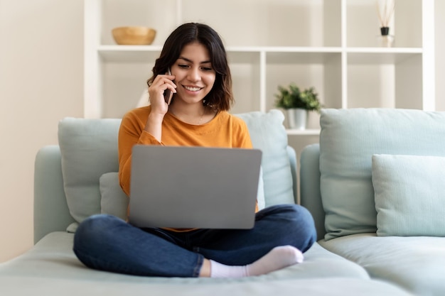 Mujer de Oriente Medio usando una computadora portátil y hablando por teléfono móvil en casa