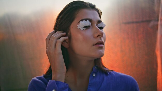 Foto mujer original tocando el cabello posando con pestañas de pétalos en un primer plano de brillo al atardecer