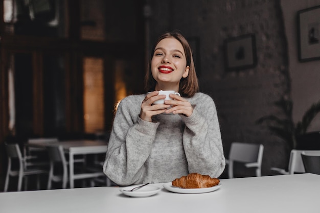 Mujer ositiva con sonrisa blanca como la nieve y lápiz labial rojo sostiene una taza de té mientras se sienta en un pequeño café