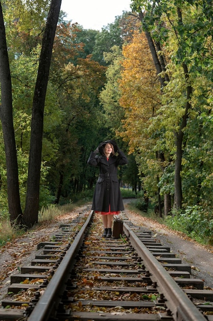 Mujer en otoño bosque ropa retro abrigo negro y sombrero de ala ancha con maleta vieja sobre rieles Mujer vestida con estilo años 40