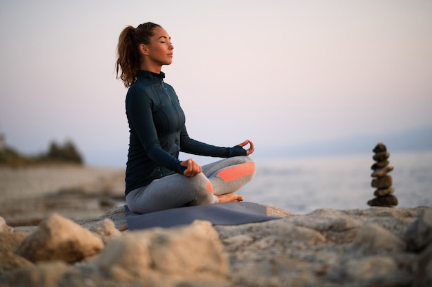 Mujer pacífica con los ojos cerrados sentada en posición de loto mientras practica Yoga en la playa rocosa.