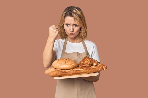 Mujer panadera caucásica de mediana edad con bandeja de pan mostrando el puño a la cámara expresión facial agresiva