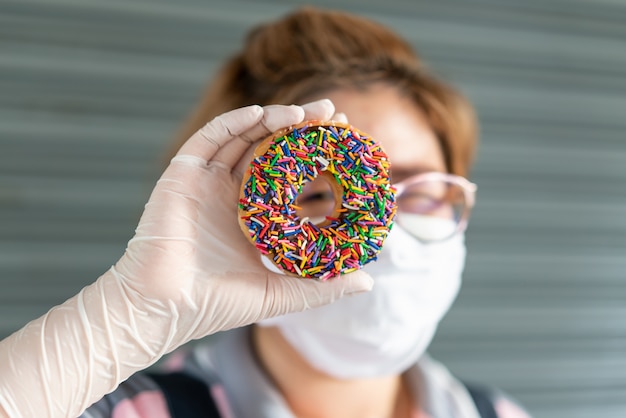 Mujer panadero con guantes de látex, sosteniendo una rosquilla helada de chocolate fresco y mirando el agujero de la rosquilla.