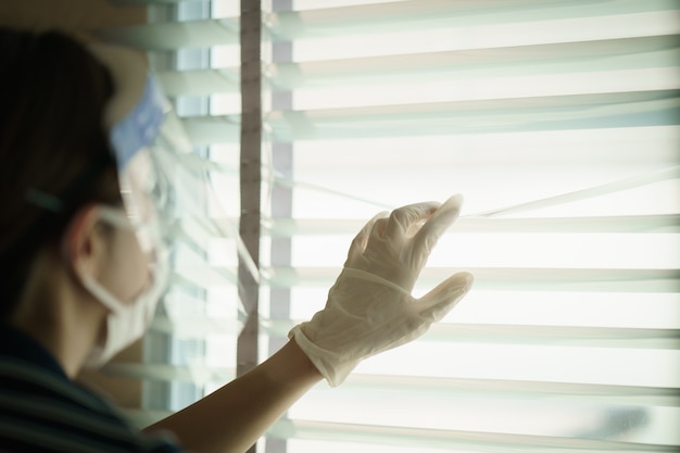 Mujer con pantalla médica de protección o protector facial de plástico, mirando a través de las persianas. Para virus corona o protección Covid-19