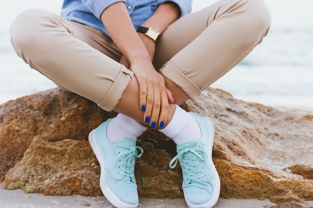 Mujer con pantalones beige y una camisa de mezclilla y zapatillas turquesas sentada en una roca junto al mar