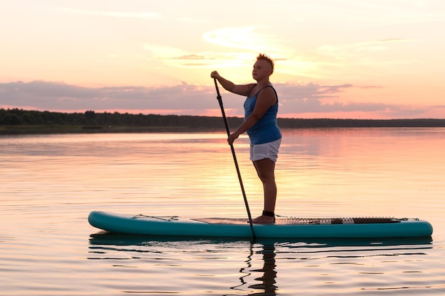 Una mujer con pantalones cortos mohawk se para en una tabla de SUP al atardecer en un lago contra un cielo azul rosa y agua