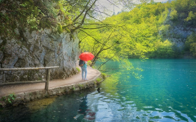 Foto mujer con paraguas rojo en el camino cerca del hermoso lago bajo la lluvia en primavera lagos de plitvice croacia paisaje colorido con sendero de niña árboles verdes roca agua azul en el parque nacional en verano pasarela