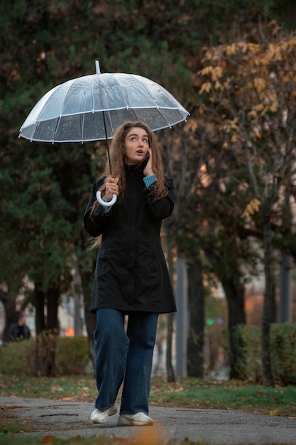 Mujer con paraguas transparente caminando en el parque en otoño y hablando por teléfono Mujer hermosa joven usando teléfono inteligente bajo paraguas