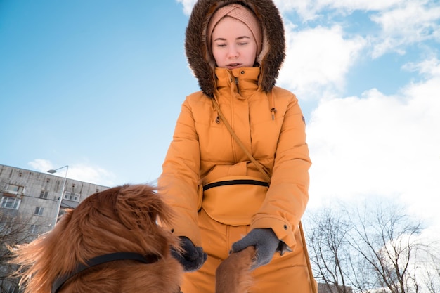 Mujer en parka amarilla con capucha de piel caminando y jugando con perro en la ciudad en día de invierno