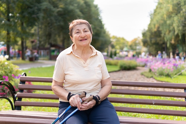 Una mujer en el parque camina nórdico con palos en un día soleado de verano. La mujer mayor de buen humor sentada en el banco.