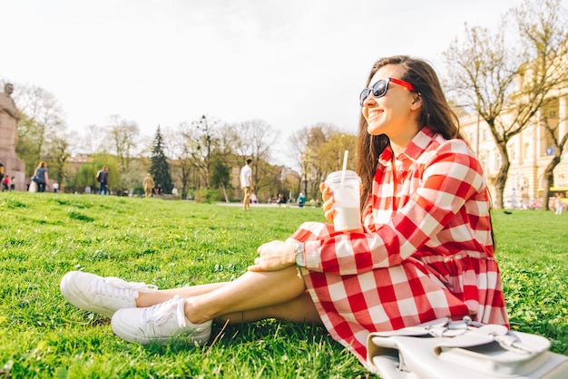 Mujer en el parque de la ciudad sentada en el suelo y bebiendo batido